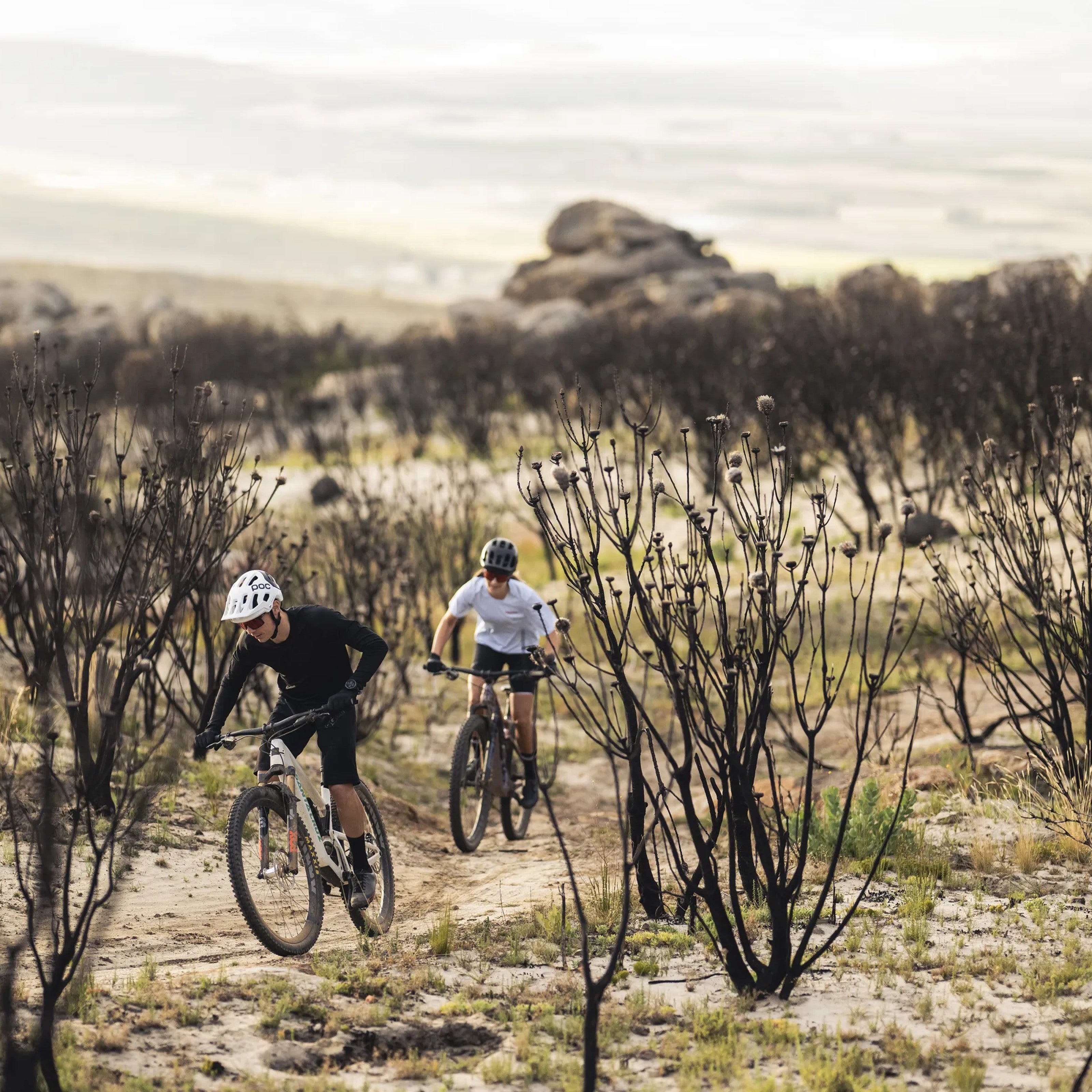 Two mountain bikers using SQLab 710 ergonomic grips, riding on a trail in a desert landscape with sparse vegetation.