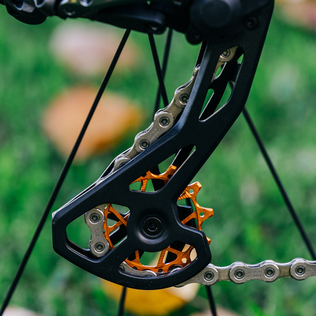 Close-up of colourful orange Garbaruk rear derailleur pulleys for Shimano mounted on custom bike, showcasing CNC-machined detail and precision engineering, photographed outdoors against a leafy green background