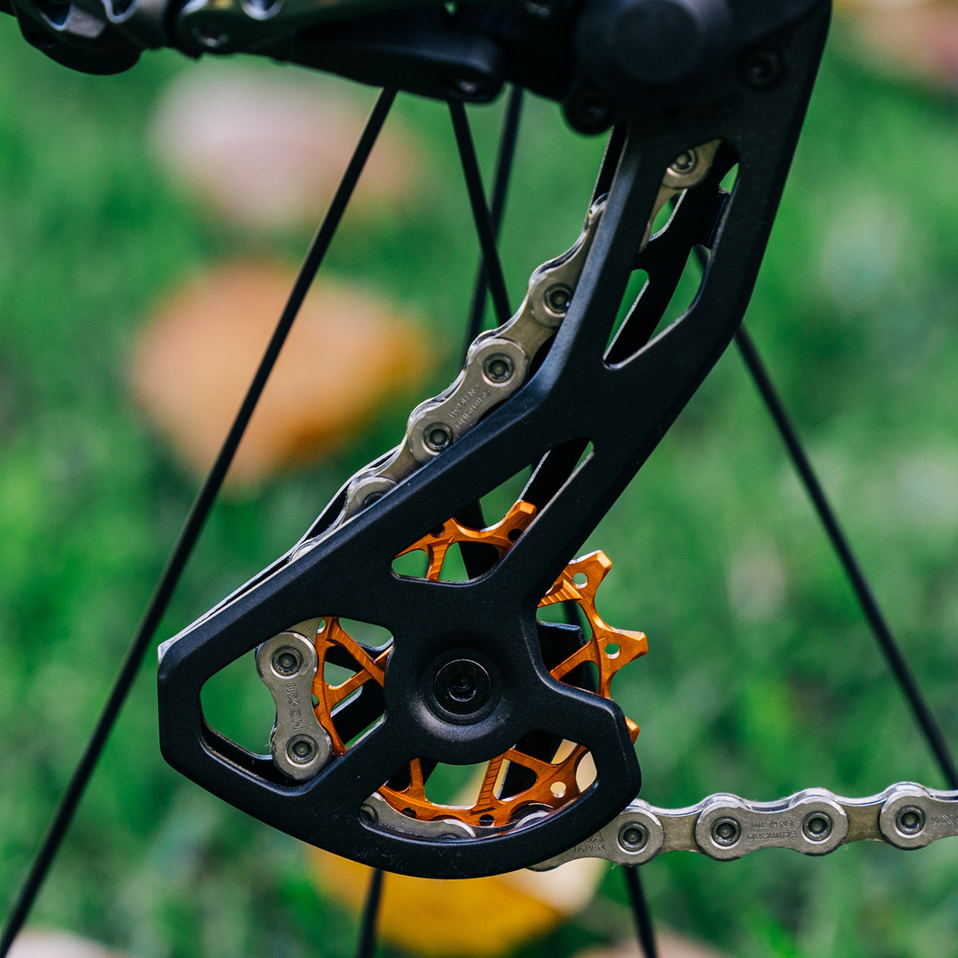 Close-up of colourful orange Garbaruk rear derailleur pulleys for Shimano mounted on custom bike, showcasing CNC-machined detail and precision engineering, photographed outdoors against a leafy green background